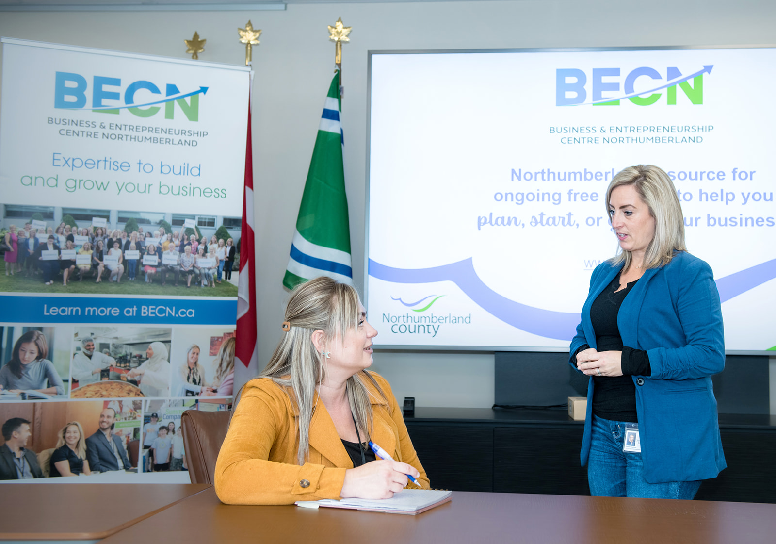 Two women at table signing document with BECN banners in background