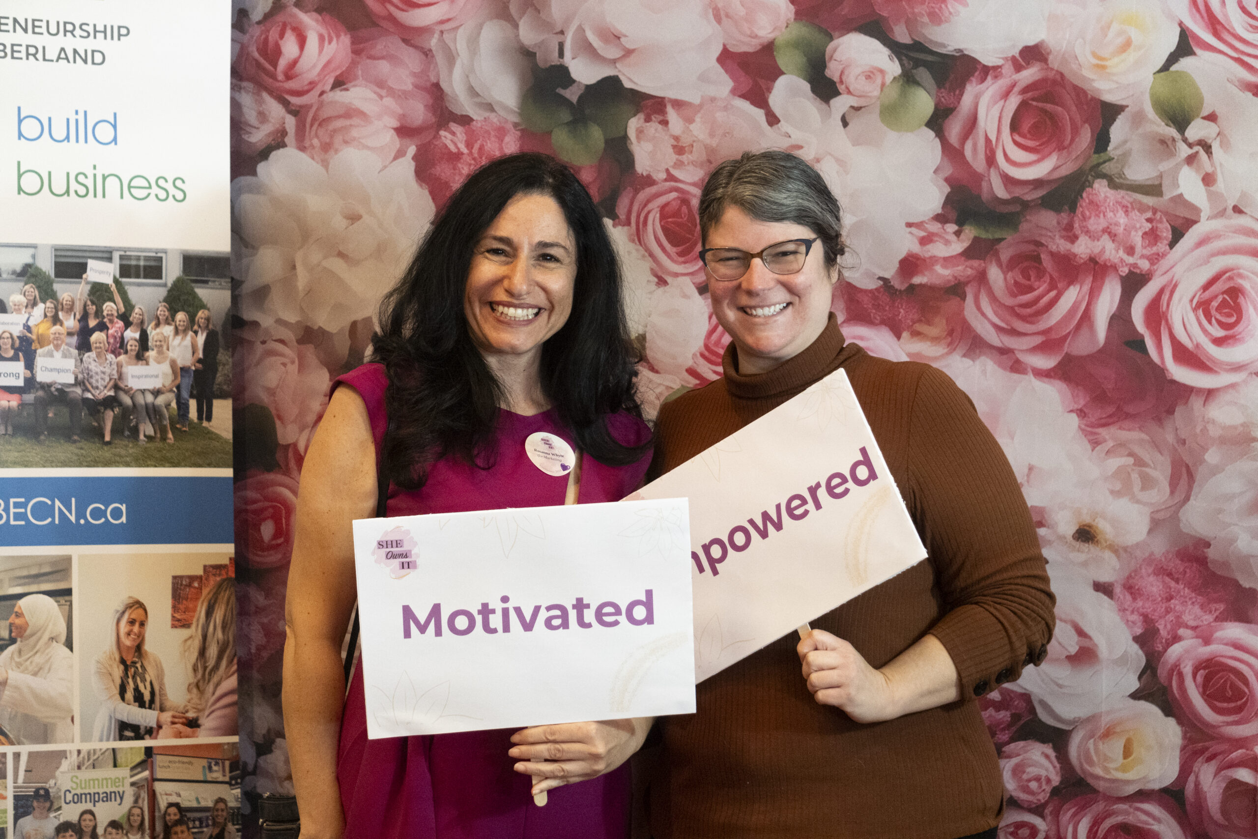 Two women smiling and holding 'Motivated' sign in front of floral backdrop